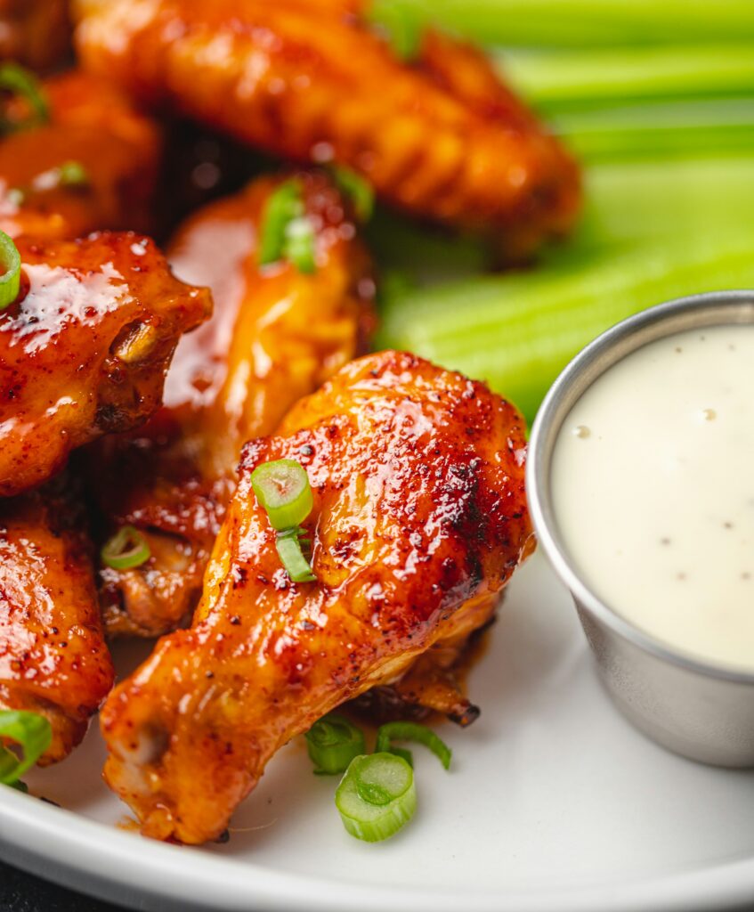 Glazed chicken wings with green onions served on a white plate with celery sticks and a small cup of ranch dipping sauce