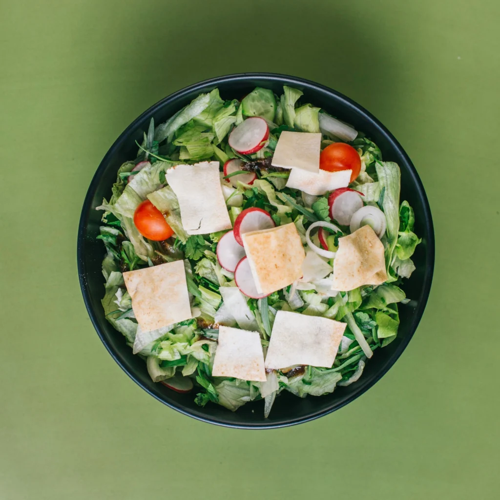 Fresh garden salad in a black bowl with lettuce, radish slices, cherry tomatoes, onion rings, cucumber, and crispy pita chips on a green background.